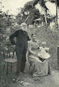 Heinrich Bohner mit weißem Rauschebart und im Anzug steht vor einem Korbstuhl, seinen Arm um eine sitzende afrikanische Frau im Kleid mit ebenfalls im Kleid gekleideten Kleinkind auf dem Schoß. Im Hintergrund Gebäude und die Landschaft Kameruns-