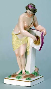 Porzellanfigur einer halbnackten Nymphe, sich ein Tuch um den Körper wickelnd an einen Pfeiler gelehnt.