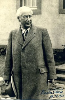 Photographie des alten Theodor Heuss, vor einem Gebäude, in Anzug und Mantel.