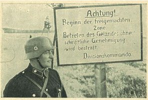 Photographie eines Soldaten im Stahlhelm neben einem Schild, darauf steht: Achtung! Beginn der freigemachten Zone, Betreten des Geländes ohne schriftliche Genehmigung wird bestraft. Divisionskommando