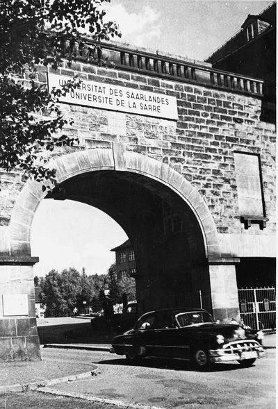 Schwarz-weiß Photographie eines Tores mit der Aufschrift Universität des Saarlandes, durch das ein Auto aus 1950er/60er Jahre fährt.