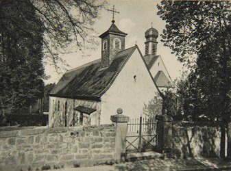 Eine Schwarzweißaufnahme einer kleinen, weißen Kapelle mit einem kleinen, bekreuzten Tümchen. Dahinter dem Gebäude steht noch eine kleine Kapelle mit einer Art Zwiebeltürmchen. Vorne im Bild ist eine Mauer und ein Eisentor.