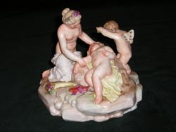 Porzellanfigur, auf einem Stein liegt ein niedergestrecktes Kind umsorgt von einer halbnackten Frau, im Hintergrund ein Eros der sich den Arm vors Gesicht hält.